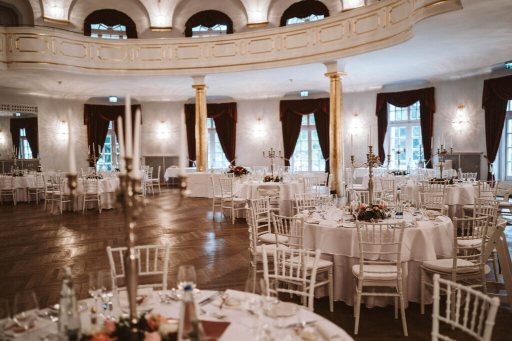 Eleganter Ballsaal im Kurhaus in Bad Tölz, eingedeckt für eine Veranstaltung mit runden Tischen mit weißen Tischdecken, floralen Tafelaufsätzen, weißen Stühlen, Kandelabern und großen, von roten Vorhängen umrahmten Fenstern unter einem Balkon mit goldenen Akzenten.