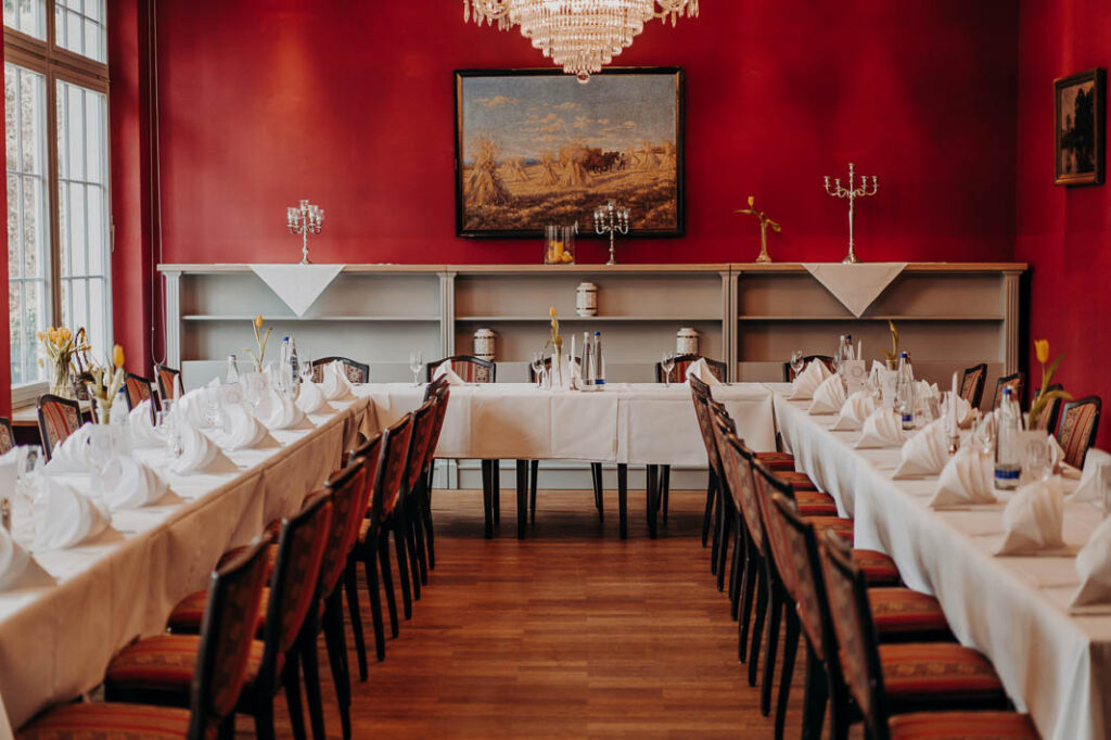 Zwei lange, formelle Esstische sind mit weißen Tischtüchern, Servietten und Gläsern in einem eleganten Raum mit roten Wänden im Kurhaus in Bad Tölz gedeckt, der mit Kronleuchtern, Kunst an den Wänden und gepolsterten Stühlen an beiden Tischen geschmückt ist.