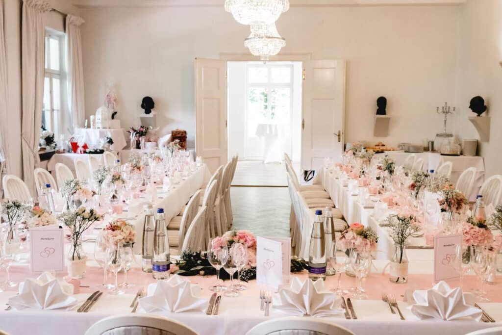 Eleganter Hochzeitssaal im Kurhaus in Bad Tölz mit zwei langen Tischen, die mit weißer Bettwäsche, rosafarbenen Blumendekorationen, gefalteten Servietten, Speisekarten und Wasserflaschen unter Kristalllüstern in einem hellen, luftigen Raum gedeckt sind.