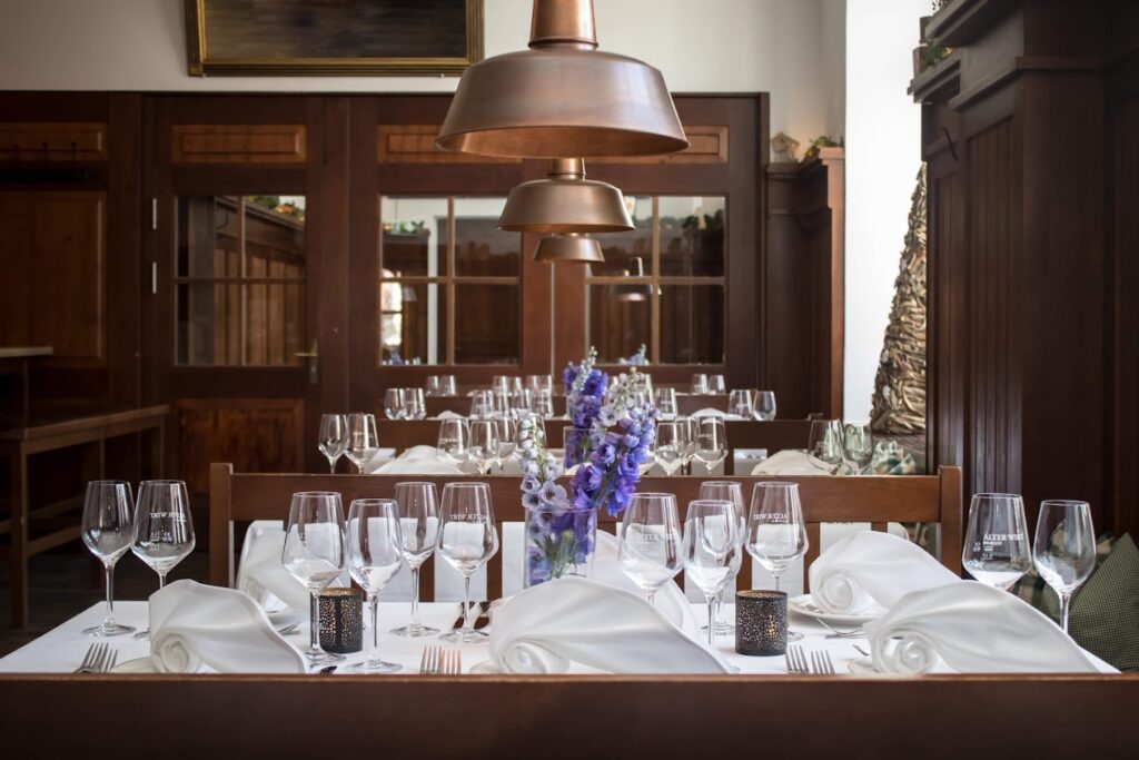 Ein fein säuberlich gedeckter Restauranttisch mit weißen Servietten, leeren Weingläsern, lila Blumen in einer Vase und Holzstühlen steht unter kupfernen Hängelampen in einem warm beleuchteten Esszimmer - eine Umgebung, in der Florian Otto seine Zauberkunst zum Leben erwecken könnte.