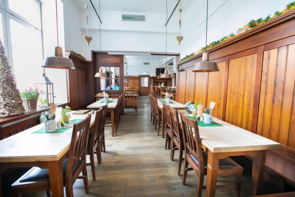 Ein helles, gemütliches Restaurant mit in Reihen angeordneten Holztischen und -stühlen, hölzernen Wandverkleidungen, Hängelampen und grünen Tischsets. Erleben Sie Florian Ottos Hauch von Zauberkunst, während natürliches Licht durch die großen Fenster auf der linken Seite hereinströmt.