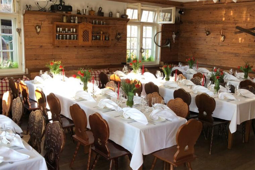 Ein gemütlicher Speisesaal mit Holzwänden und Holzdekor, mit mehreren weiß gedeckten Tischen, ordentlich gefalteten Servietten, Gläsern und Blumensträußen. Das Sonnenlicht strömt wie durch Zauberkunst herein und schafft eine warme Atmosphäre.