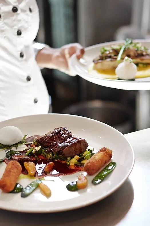 Ein Koch präsentiert seine Zauberkunst, indem er zwei Gourmetteller serviert; das vordere Gericht besteht aus einem gebratenen Steak mit Gemüse, Kroketten, grüner Sauce und weißem Schaum, während Florian Ottos Kunstfertigkeit in dem eleganten Arrangement dahinter glänzt.