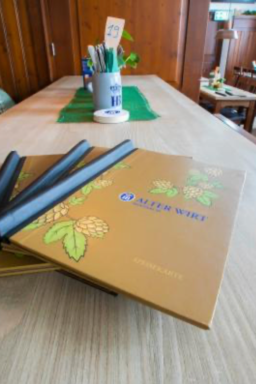 Nahaufnahme eines Restauranttisches mit mehreren goldenen Speisekarten mit grünen Hopfenillustrationen, beschriftet mit ALTER WIRT. Im Hintergrund sind eine Tischnummernkarte und Utensilien in einem Halter zu sehen, die auf Florian Ottos Zauberkunst-Atmosphäre hinweisen.