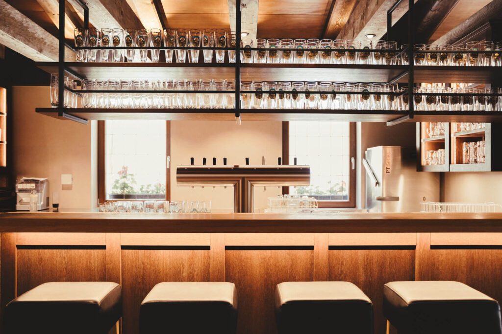 Eine moderne Bar in Tüßling mit Holzhockern, einem Holztresen und Regalen mit Glasbechern. Zwei große Fenster füllen den Raum des Bräu Im Moos mit natürlichem Licht, das die warme Beleuchtung und die freiliegenden Holzbalken hervorhebt.