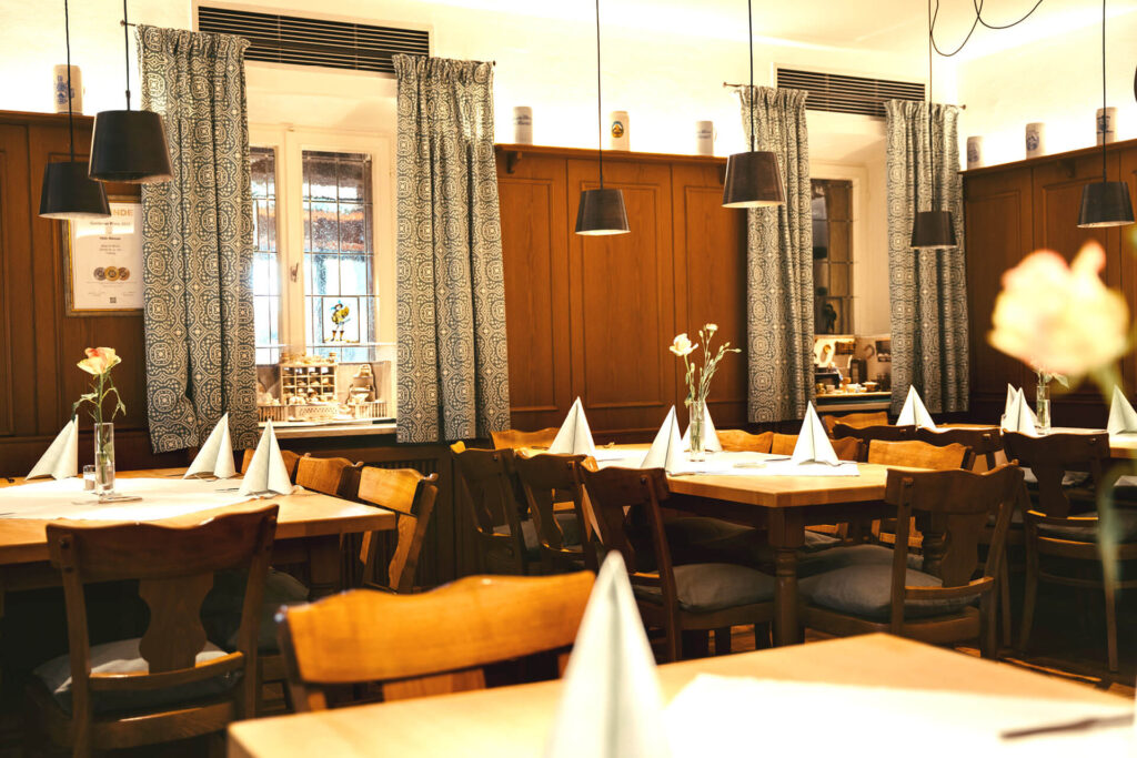 Ein gemütlicher Restaurantbereich im Bräu Im Moos in Tüßling mit Holztischen und -stühlen, weißen Servietten, die auf jedem Tisch gefaltet sind, einzelnen Blumenvasen, Hängeleuchten und gemusterten Vorhängen an den Fenstern.