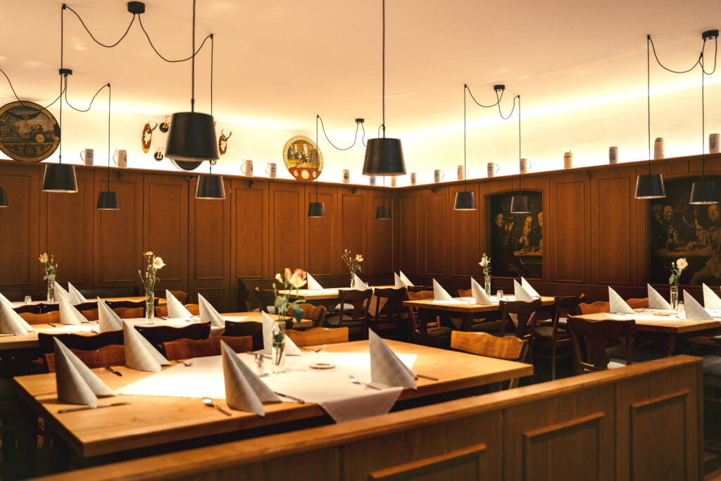 Ein gemütlicher Speisesaal im Bräu Im Moos in Tüßling ist mit Holztischen und -stühlen, weißen gefalteten Servietten und Blumen ausgestattet. Schwarze Hängelampen hängen von der Decke, während dekorative Teller und Kunstwerke die holzgetäfelten Wände säumen.