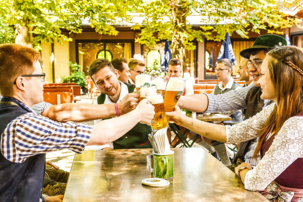 Eine Gruppe von Menschen sitzt an einem Tisch im Freien im Biergarten des Bräu Im Moos in Tüßling, lächelt und stößt unter Laubbäumen mit großen Biergläsern an.