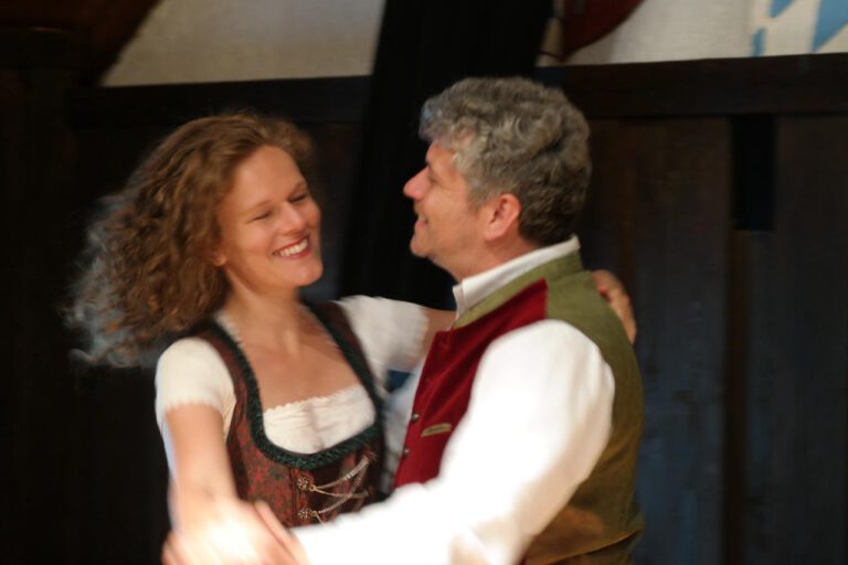 Eine lächelnde Frau und ein lächelnder Mann in traditioneller bayerischer Kleidung tanzen zusammen in einer Halle. Der dunkle Hintergrund hebt ihre freudigen Gesichtsausdrücke und lebhaften Bewegungen hervor und fängt die festliche Stimmung einer Veranstaltung des Comedydinner München ein.