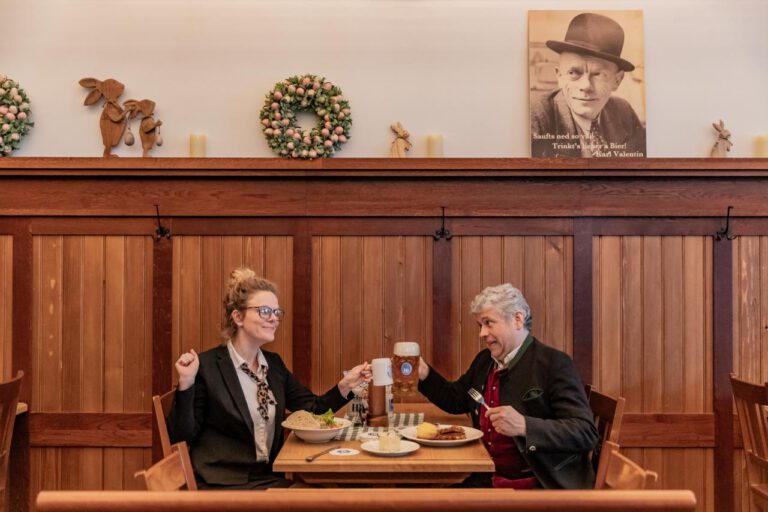 Zwei Personen sitzen an einem Holztisch in einem gemütlichen Restaurant in München und stoßen mit Getränken auf das Essen an. An der Wand hinter ihnen hängen Kränze, dekorative Figuren und ein Porträt - wie geschaffen für einen Comedydinner-Abend.