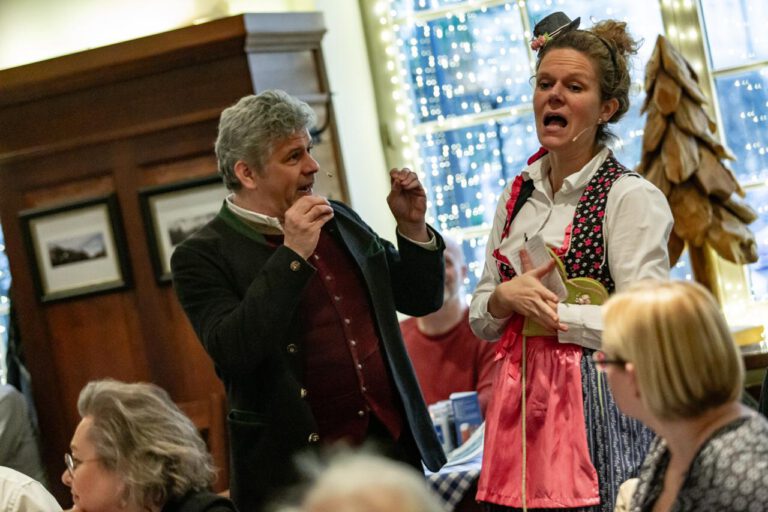 Ein Mann und eine Frau in traditioneller bayerischer Kleidung singen oder spielen zusammen bei einer lebhaften Comedydinner München Veranstaltung, umgeben von sitzenden Gästen. Der gemütliche Innenbereich ist mit gerahmten Fotos und Lichterketten dekoriert.