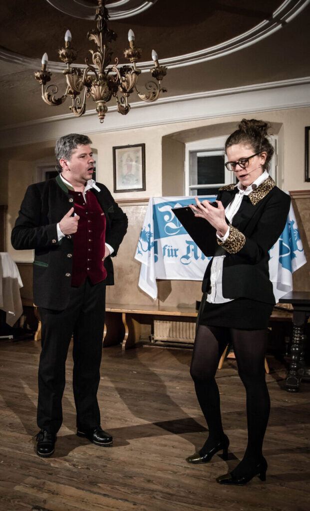 Ein Mann in formeller Kleidung im älteren Stil spricht mit einer Frau in einer schwarz-goldenen Jacke, die von einem Klemmbrett abliest. In einem Innenraum unter einem Kronleuchter stehen sie vor einem weiß-blauen Comedydinner München-Banner im Hintergrund.