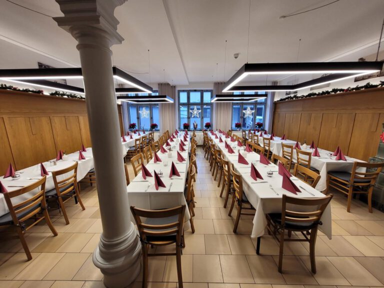 Im Gasthof zur Post in Frontenhausen stehen lange Tische mit weißen Tischtüchern, Holzstühlen und roten Servietten bereit für ein Festessen. Große Fenster und festliche Dekorationen verstärken das einladende Ambiente.