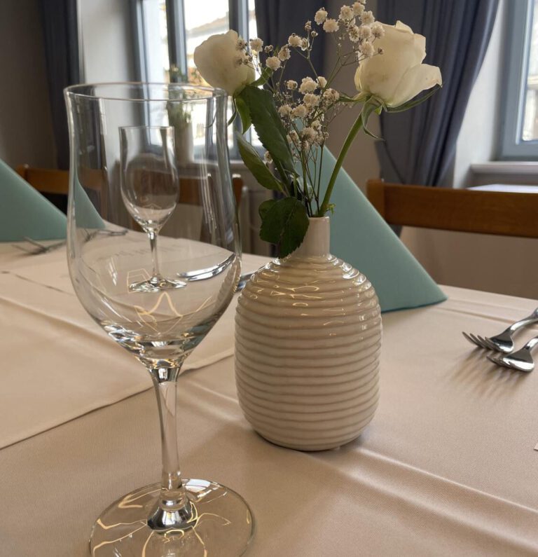 Nahaufnahme eines ordentlich gedeckten Esstisches im Gasthof zur Post in Frontenhausen mit einem Weinglas, einer gestreiften Keramikvase mit weißen Blumen, gefalteten blauen Servietten und Besteck in einem hellen Raum mit großen Fenstern.