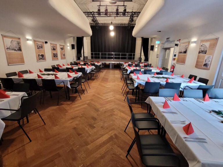 In einem Festsaal des Gasthofs zur Post in Frontenhausen stehen lange Tische mit weißen Tüchern und roten Servietten, polierten Utensilien und Stühlen. Gerahmte Fotos säumen die Wände, und am anderen Ende steht eine Bühne mit Scheinwerferlicht.