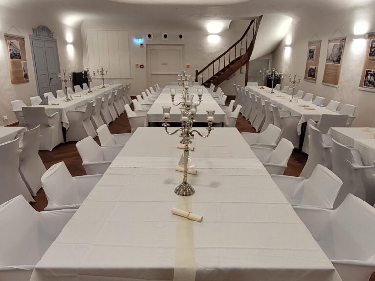 Im Festsaal des Gasthofs zur Post in Frontenhausen sind die mit weißen Tüchern gedeckten Tische, umgeben von weißen Stühlen, fein säuberlich angeordnet. Silberne Kandelaber schmücken die Tische, gerahmte Bilder säumen die Wände, und eine Holztreppe ziert den Hintergrund.