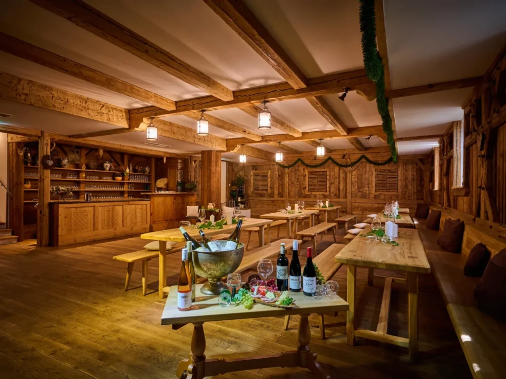 Im Happinger Hof in Rosenheim steht ein gemütlicher hölzerner Speisesaal mit langen, mit Girlanden geschmückten Tischen und Bänken. Flaschen, Gläser und Eiskübel stehen auf dem vorderen Tisch, während die warme Beleuchtung eine einladende, rustikale Atmosphäre schafft.