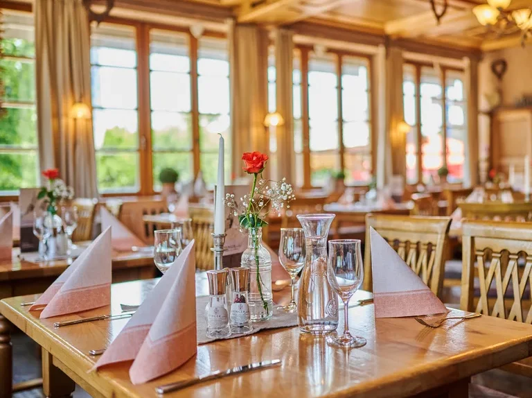 Der helle, elegante Speisesaal des Happinger Hofs in Rosenheim ist mit ordentlich gedeckten Holztischen, gefalteten Servietten, Gläsern, Besteck und kleinen Vasen mit roten Rosen ausgestattet; große Fenster lassen viel natürliches Licht in den Raum.