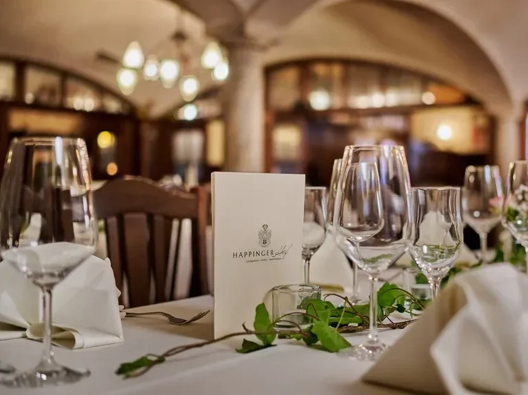 Nahaufnahme eines elegant gedeckten Tisches im Happinger Hof in Rosenheim mit Weingläsern, weißen Servietten, einer Speisekarte und grüner Blattdekoration, mit einem sanft beleuchteten, gewölbten Speisesaal im Hintergrund.