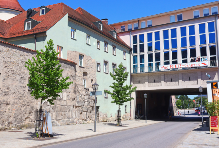 Eine ruhige Straße in Regensburg mit pastellfarbenen Gebäuden, alten Steinmauern und jungen Bäumen. Ein Torbogen verbindet zwei Gebäude mit einem ProBIER-Garten-Schild über dem Durchgang und dem Kolpinghaus in der Nähe.
