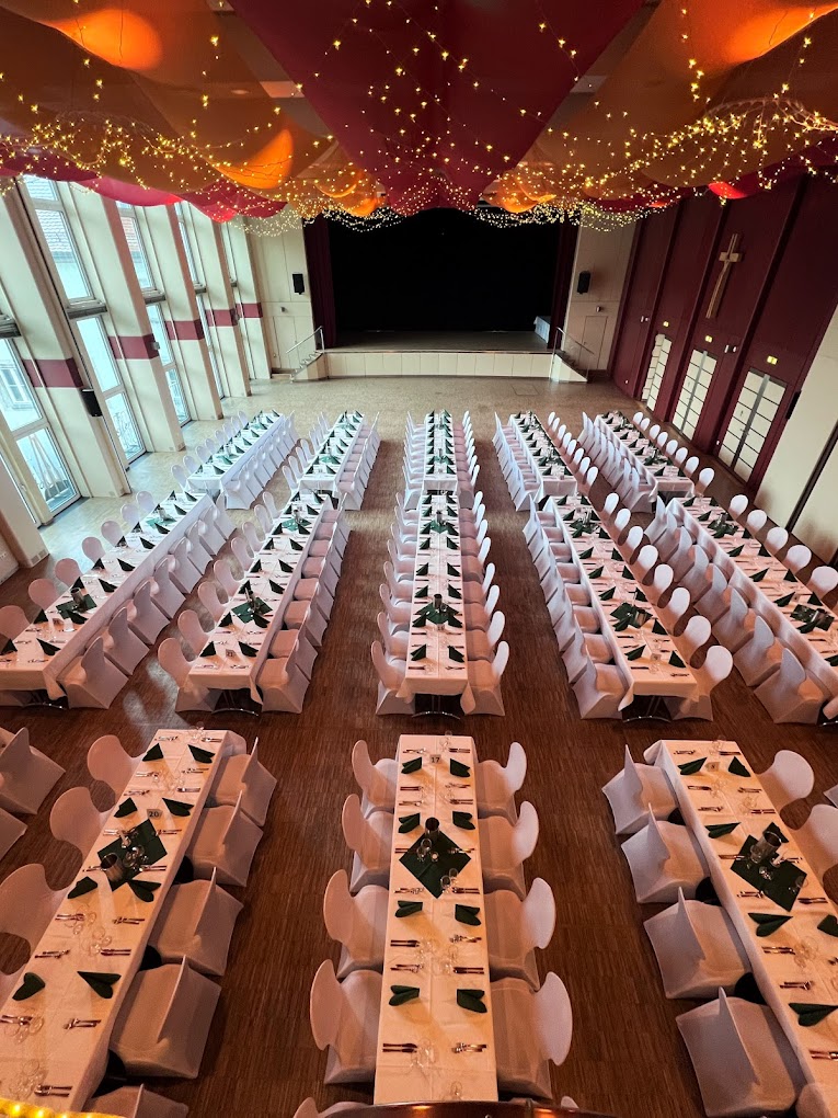 In einem großen Festsaal des Kolpinghauses in Regensburg stehen lange Tischreihen mit weißen Tischdecken und Stühlen. Warme Lichterketten und drapierte Stoffe schmücken die Decke und schaffen eine festliche Atmosphäre, die für jede Veranstaltung geeignet ist.