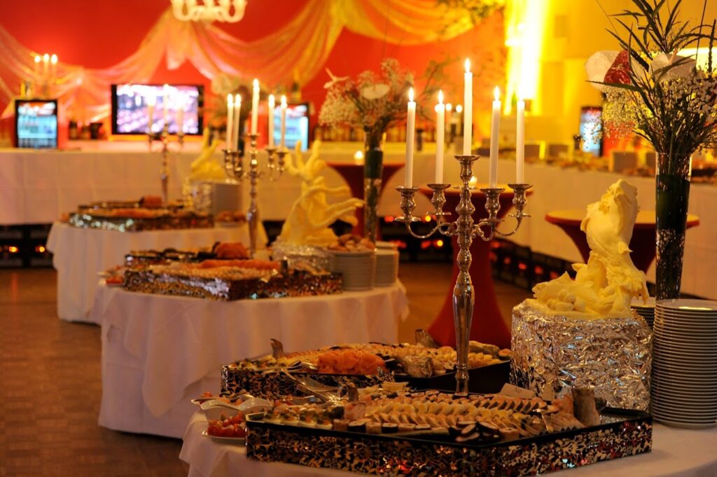 Ein üppiger Buffetaufbau im Kolpinghaus Regensburg, warm beleuchtet mit Tischen mit verschiedenen Gerichten, Kandelabern, dekorativen Blumen und eleganten, mit Folie überzogenen Speisenauslagen. Im Hintergrund sind drapierte Stoffe und leuchtende Lichter zu sehen.