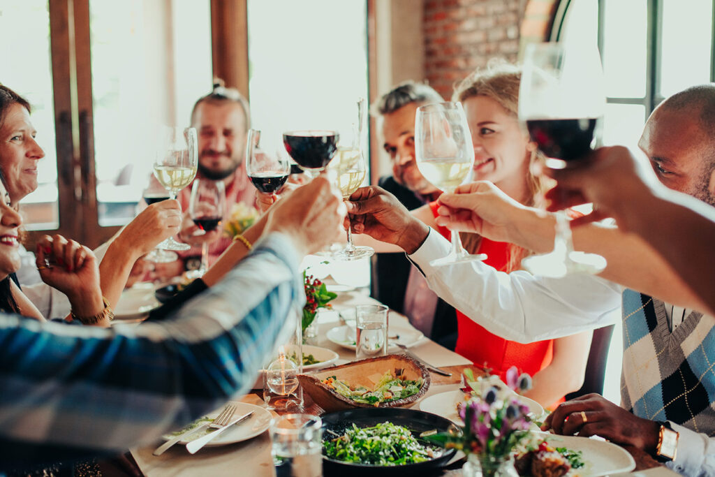 Eine Gruppe von Menschen sitzt bei einem Firmenfeier-Krimidinner in München um einen Tisch herum, stößt mit Gläsern an und lächelt. Der Tisch ist mit Speisen, Getränken und Blumen gedeckt und schafft eine warme, feierliche Atmosphäre.