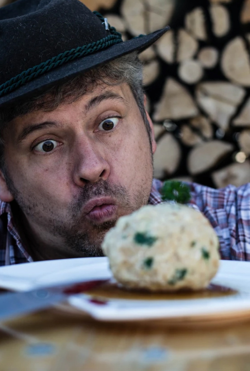 Ein Mann mit Trachtenhut und kariertem Hemd macht ein erstauntes Gesicht, als er einen großen Knödelkomplott mit Petersilie auf einem Teller betrachtet, während im Hintergrund Brennholz aufgeschichtet ist.