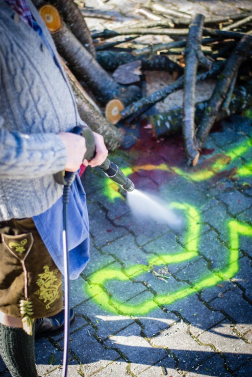 Eine Person in traditioneller Kleidung, vielleicht inspiriert durch das Knödelkomplott, reinigt mit einem Hochdruckreiniger ein mit grüner Sprühfarbe markiertes Steinpflaster, während im Hintergrund abgeschnittene Äste aufgestapelt werden.