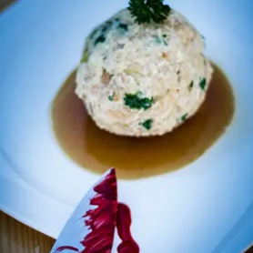 Ein runder Knödelkomplott, garniert mit Petersilie, liegt in einer flachen Pfütze aus brauner Soße auf einem weißen Teller. Ein Messer mit rot-weißem Griff liegt daneben.