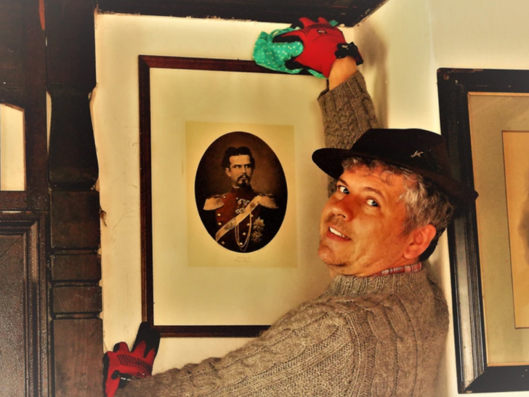 Ein Mann mit braunem Hut, roten Handschuhen und beigem Pullover lächelt in die Kamera, während er die obere Ecke eines gerahmten Porträts eines bärtigen Mannes in Uniform säubert - eine Szene, die einem Knödelkomplott-Krimi würdig ist.