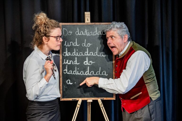 Eine Frau mit Brille steht neben einer Kreidetafel mit verspielter Schrift, während ein älterer Mann in Weste und Fliege ausdrucksstark auf die Tafel zeigt - die Kulisse für ein lebendiges Krimidinner München. Im Hintergrund hängen schwarze Vorhänge.