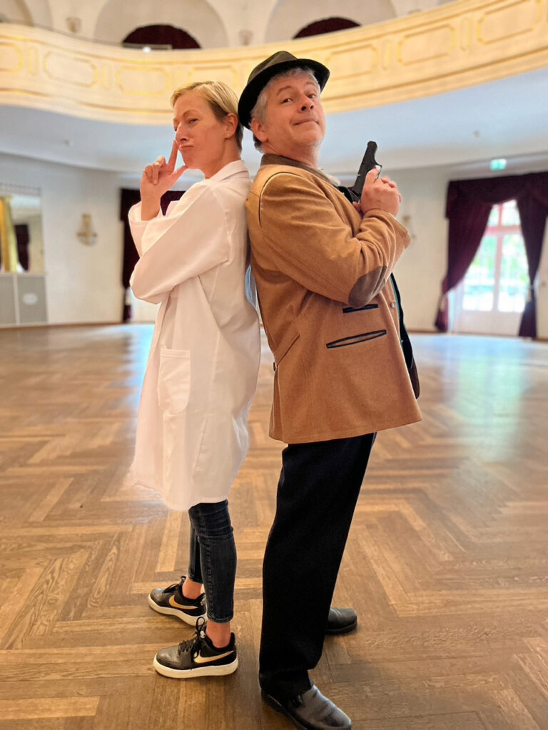 Zwei Erwachsene stehen Rücken an Rücken in einem großen, eleganten Raum. Der eine trägt einen weißen Laborkittel und Turnschuhe, der andere eine braune Jacke, einen schwarzen Hut und eine Pistolenrequisite - und wirft sich beim Krimidinner in München in Pose.