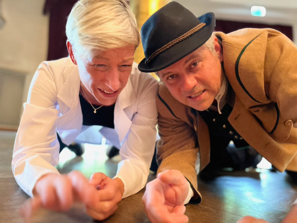 Zwei Erwachsene, einer in einem weißen Mantel und der andere in traditioneller brauner Kleidung mit Hut, liegen auf einem Holzboden in einem Innenraum, lächeln und strecken sich der Kamera entgegen. Die verspielte Szene fängt die fröhliche Stimmung eines Krimidinners in München ein.