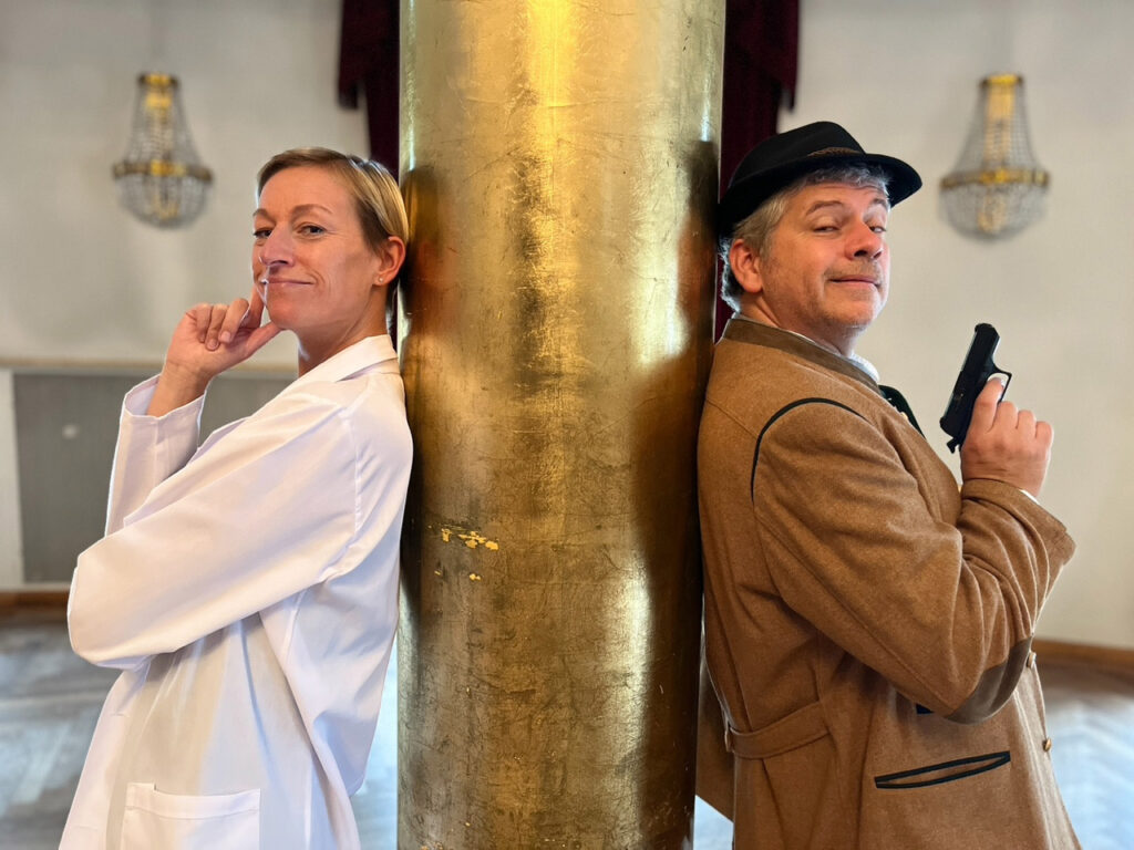 Eine Frau im weißen Laborkittel und ein Mann mit braunem Mantel und Mütze, der eine Spielzeugpistole in der Hand hält, stehen Rücken an Rücken an einer goldenen Säule, beide lächeln verschmitzt - bereit für ihr nächstes Krimidinner in München-Abenteuer.