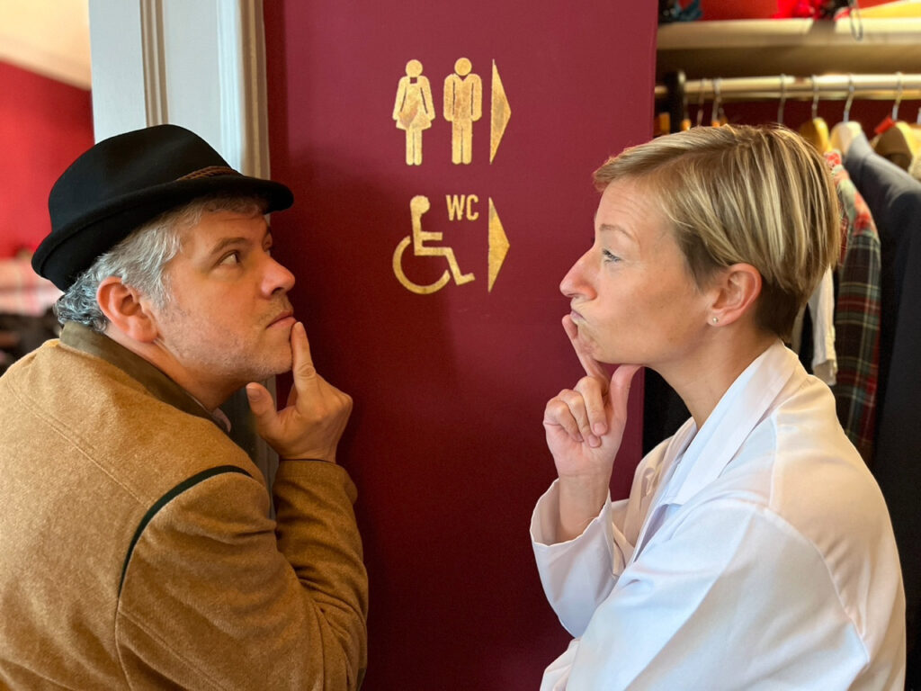Ein Mann und eine Frau stehen an einer kastanienbraunen Wand mit Toilettenschildern, beide blicken nachdenklich mit einem Finger am Kinn; es scheint, als würden sie bei einem Krimidinner in München über Hinweise nachdenken, während im Hintergrund Kleidung hängt.