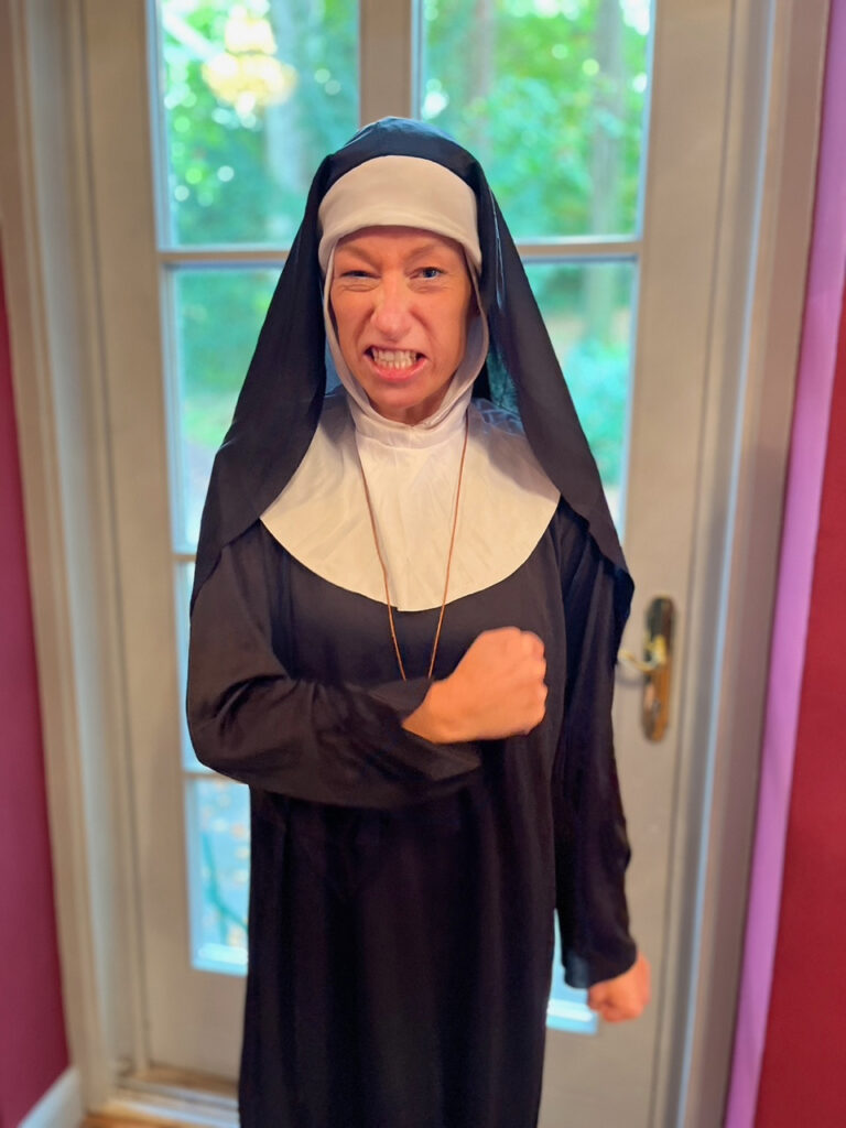 Eine in ein Nonnenkostüm gekleidete Person steht drinnen, macht ein spielerisch wütendes Gesicht und ballt die Faust, als ob sie sich auf ein Krimidinner in München vorbereiten würde, wobei durch die Glastür hinter ihr Grünzeug zu sehen ist.