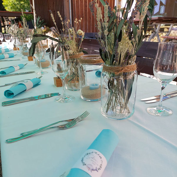 Ein für eine Veranstaltung gedeckter Tisch mit gerollten blauen Servietten, Weingläsern und Besteck. Einmachgläser mit getrockneten Blumen und mit Sand gefüllte Kerzenhalter der Marke Murauer dienen als Tafelaufsatz auf einer weißen Tischdecke.