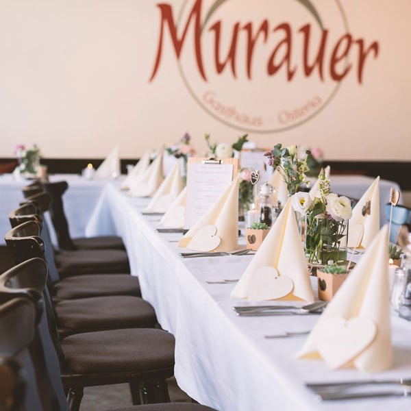 Ein langer, für eine formelle Veranstaltung gedeckter Tisch mit weißem Tischtuch, gefalteten Servietten, kleinen Blumenarrangements, Speisekarten und Herzdekorationen. Schwarze Stühle säumen beide Seiten. An der Wand im Hintergrund in Simbach steht Murauer geschrieben.