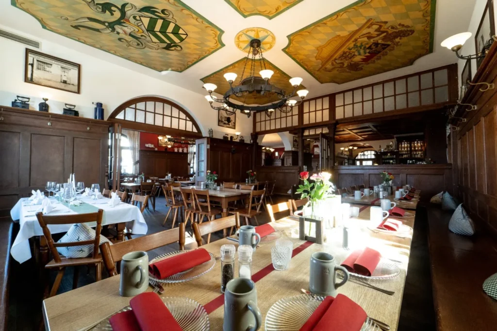 Ein traditioneller Speisesaal in München mit Holzwänden, einer verzierten Decke und einem großen Kronleuchter. Die Tische im Haderner Augustiner sind mit roten Servietten, Tellern, Gläsern und Keramikbechern gedeckt. Sonnenlicht strömt herein und schafft eine warme Atmosphäre.