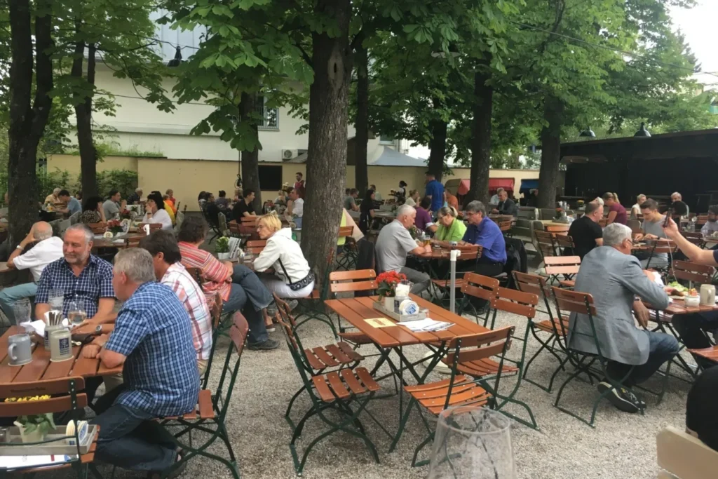 In einem Biergarten in München sitzen Menschen an Holztischen und Bänken im Freien, unterhalten sich und genießen Haderner Augustiner unter grünen Bäumen. Die Szene ist lebendig und schattig, mit viel Grün und einer entspannten Atmosphäre.