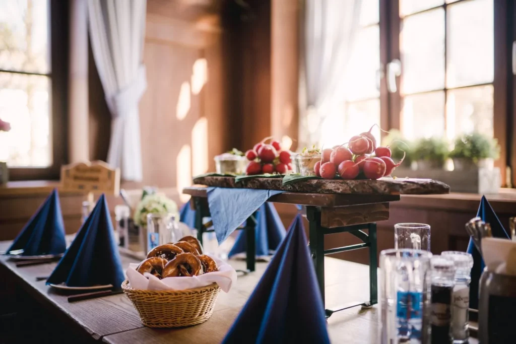 Ein rustikaler Holztisch ist mit blauen, zu Dreiecken gefalteten Servietten, einem Korb mit Brezeln, Gläsern und Tabletts mit Vorspeisen - darunter Radieschen - gedeckt und lädt dazu ein, zusammen mit Haderner Augustiner einen Hauch von München zu genießen. Durch die weiß getäfelten Fenster strömt das Sonnenlicht herein.